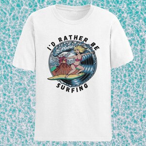 Puede incluir: Camiseta blanca con un gr&aacute;fico de una mujer surfeando una ola con un volc&aacute;n en erupci&oacute;n en el fondo. El texto "I'd Rather Be Surfing" est&aacute; impreso en un c&iacute;rculo alrededor del gr&aacute;fico.
