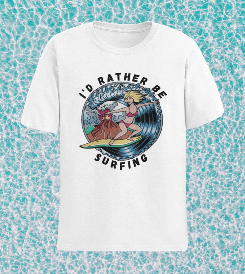 Puede incluir: Camiseta blanca con un gr&aacute;fico de una mujer surfeando una ola con un volc&aacute;n en erupci&oacute;n en el fondo. El texto "I'd Rather Be Surfing" est&aacute; impreso en un c&iacute;rculo alrededor del gr&aacute;fico.