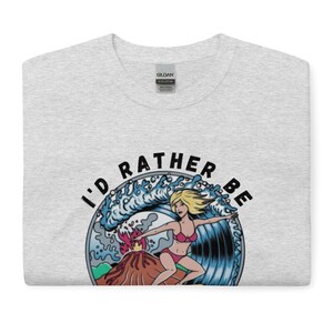 Puede incluir: Una camiseta gris con un gr&aacute;fico de una mujer surfeando una ola con un volc&aacute;n en erupci&oacute;n en el fondo. El texto "I'd rather be" est&aacute; escrito alrededor del gr&aacute;fico.
