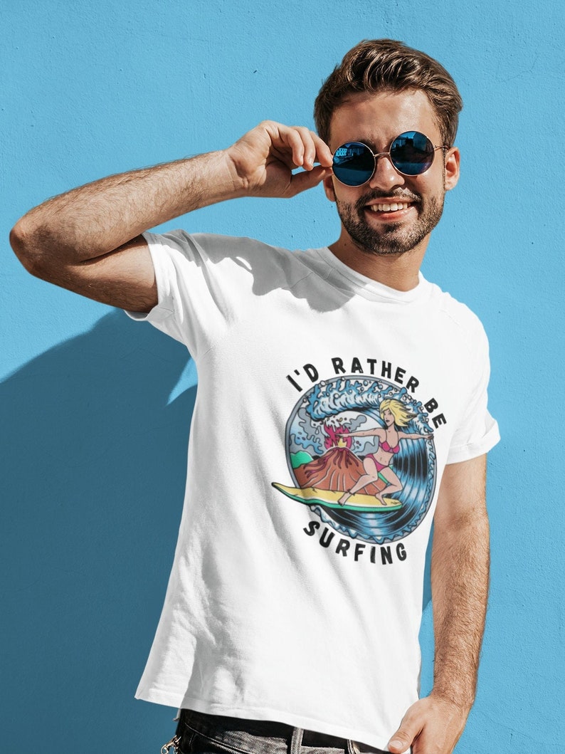 Puede incluir: Camiseta blanca con un gr&aacute;fico de una mujer surfeando en una ola con un volc&aacute;n en el fondo. El texto "I'd Rather Be Surfing" est&aacute; impreso en un c&iacute;rculo alrededor del gr&aacute;fico.