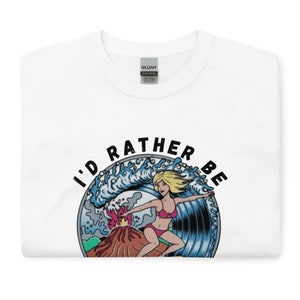 Puede incluir: Camiseta blanca con un gr&aacute;fico de una mujer surfeando una ola con un volc&aacute;n en erupci&oacute;n en el fondo. El texto "I'd rather be" est&aacute; escrito en un c&iacute;rculo alrededor del gr&aacute;fico.