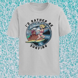 Puede incluir: Una camiseta gris claro con un gr&aacute;fico de una mujer surfeando una ola con un volc&aacute;n en erupci&oacute;n en el fondo. El texto "I'd Rather Be Surfing" est&aacute; impreso en un c&iacute;rculo alrededor del gr&aacute;fico.