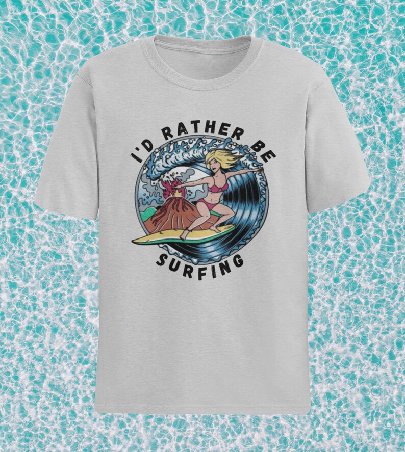 Puede incluir: Una camiseta gris claro con un gr&aacute;fico de una mujer surfeando una ola con un volc&aacute;n en erupci&oacute;n en el fondo. El texto "I'd Rather Be Surfing" est&aacute; impreso en un c&iacute;rculo alrededor del gr&aacute;fico.