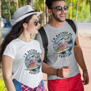 Puede incluir: Dos personas con camisetas blancas con un gr&aacute;fico de una mujer surfeando en una ola y el texto "I'd rather be surfing". La mujer del gr&aacute;fico lleva un bikini rojo y tiene el pelo rubio. Las camisetas son grises y blancas.