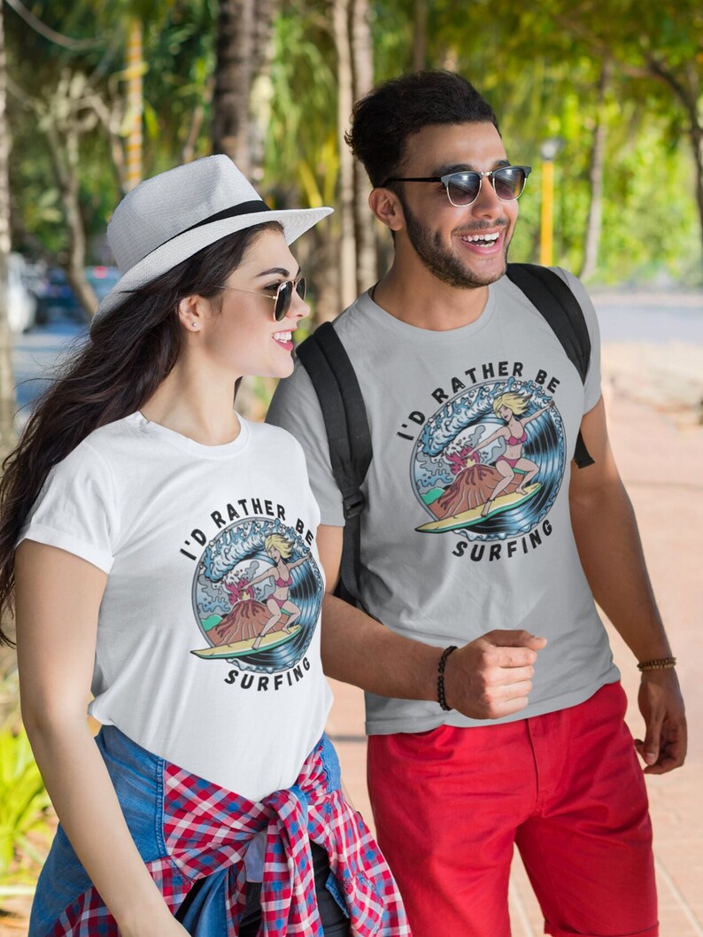 Puede incluir: Dos personas con camisetas blancas con un gr&aacute;fico de una mujer surfeando en una ola y el texto "I'd rather be surfing". La mujer del gr&aacute;fico lleva un bikini rojo y tiene el pelo rubio. Las camisetas son grises y blancas.