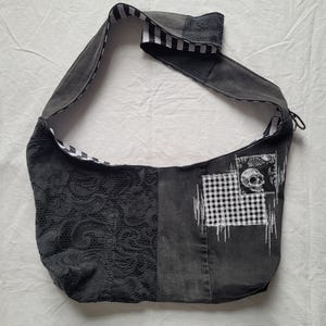 May include: A hobo-style shoulder bag crafted from dark grey denim and black lace fabric. The bag features a wide strap with a black and white striped lining. A patch on the front displays a white skull graphic over a black and white checkered pattern.