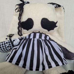 May include: A plush rabbit toy with cream-coloured fur, featuring black heart-shaped eyes and a black and white vertical striped skirt. The rabbit is adorned with black lace trim on one ear, a black and white beaded bracelet, and a black lace wristband.