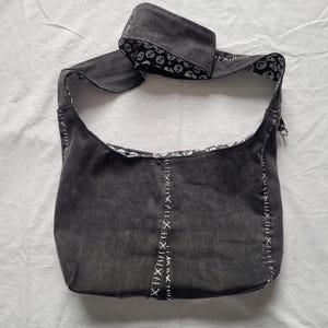May include: A dark grey denim hobo bag featuring a wide shoulder strap and a white skull pattern on the interior lining. The exterior is accented with prominent white cross-stitch detailing along the vertical seams and the bag edges.