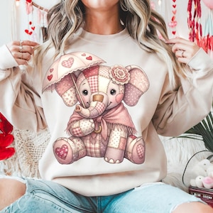 Puede incluir: Una sudadera beige con un encantador diseño de elefante de patchwork. El elefante sostiene un paraguas decorado con corazones y lleva una bufanda rosa. El diseño incorpora corazones rosas, rojos y blancos, y una flor en la oreja del elefante. El tema es el Día de San Valentín.