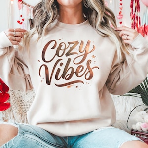 May include: A beige sweatshirt with the words "Cozy Vibes" in a copper-colored script. The sweatshirt is long-sleeved and has a relaxed fit. The image suggests a comfortable and casual style, perfect for lounging.