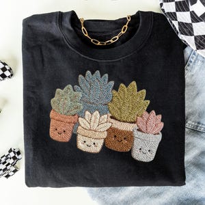 May include: Black sweatshirt featuring an embroidered design of several smiling potted succulents in various colors. A gold chain necklace is draped around the neckline. The succulents are in shades of green, blue, beige, and pink.