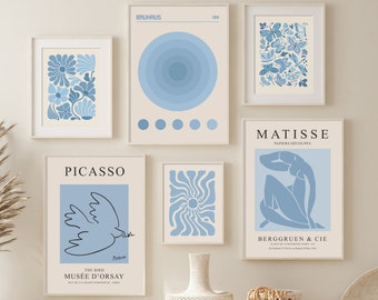 Op de afbeelding: Een set van zes blauwe en witte abstracte kunstprints met de namen van beroemde kunstenaars, waaronder Picasso, Matisse en Bauhaus. De prints zijn ingelijst en hangen aan een witte muur boven een witte bank met twee rieten manden.