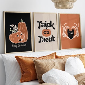 May include: Three framed Halloween-themed art prints. The first features pumpkins and the text "Happy Halloween." The second says "Trick or Treat." The third shows a black cat within a heart made of skeleton hands. The prints have a neutral color palette.