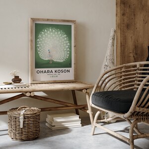 May include: A framed print of a white peacock with its tail feathers spread out. The print is on a green background and has the text "OHARA KOSON" and "PEACOCK (1945 - 1936)" in black text. The print is hanging on a white wall above a wooden bench with a wicker basket and books.