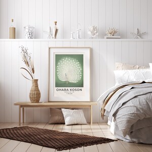 May include: A white peacock with its tail feathers spread out is featured in a vintage poster with a green background. The poster is framed and hanging on a white wall above a wooden bench. The text on the poster reads "Ohara Koson, Painter of Birds, 小原古邨".