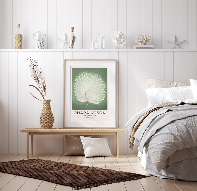 May include: A white peacock with its tail feathers spread out is featured in a vintage poster with a green background. The poster is framed and hanging on a white wall above a wooden bench. The text on the poster reads "Ohara Koson, Painter of Birds, 小原古邨".