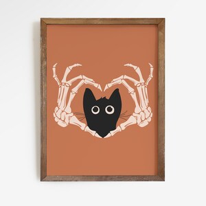 May include: A framed art print featuring a black cat head with large white eyes, framed by two beige skeleton hands forming a heart shape. The background is a solid orange color, and the frame is brown.