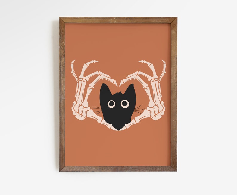 May include: A framed art print featuring a black cat head with large white eyes, framed by two beige skeleton hands forming a heart shape. The background is a solid orange color, and the frame is brown.
