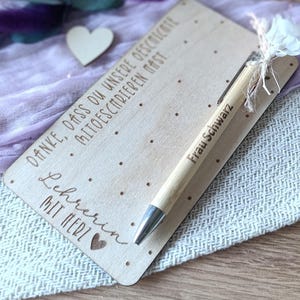 K&ouml;nnte beinhalten: Ein hellbrauner Holzstift und eine Holzkarte mit graviertem Text. Der Stift hat silberne Akzente und eine kleine wei&szlig;e Blume. Die Karte tr&auml;gt den Text "Danke, dass du unsere Geschichte mitschreiben hast. Lehrerin mit Herz" und den Namen "Frau Schwarz".