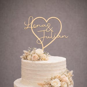 Könnte beinhalten: Zweistöckige Hochzeitstorte mit weißem Zuckerguss und Blumendekorationen. Ein herzförmiger Tortenaufsatz aus Holz mit den Namen "Lena & Julian" ist oben platziert. Die Torte steht auf einem Kuchenständer aus Holz.