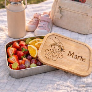 Könnte beinhalten: Eine Edelstahl-Lunchbox mit frischem Obst, einem Holzdeckel mit Marienkäfer-Design und dem Namen "Marie", einem passenden Rucksack, einer rosa Wasserflasche und rosa Turnschuhen. Ideal für ein Picknick oder ein Schulmittagessen.