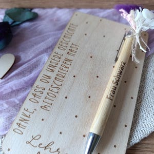 K&ouml;nnte beinhalten: Eine Holzkarte mit graviertem Text und einem Bambusstift. Der Stift ist mit "Frau Schwarz" beschriftet. Auf der Karte stehen die Worte "Danke, dass du unsere geschichte mitschreiben mast" und "Lehrerin mit Herz". Ein kleiner Strau&szlig; wei&szlig;er Blumen und lila Federn ist am Stift befestigt.