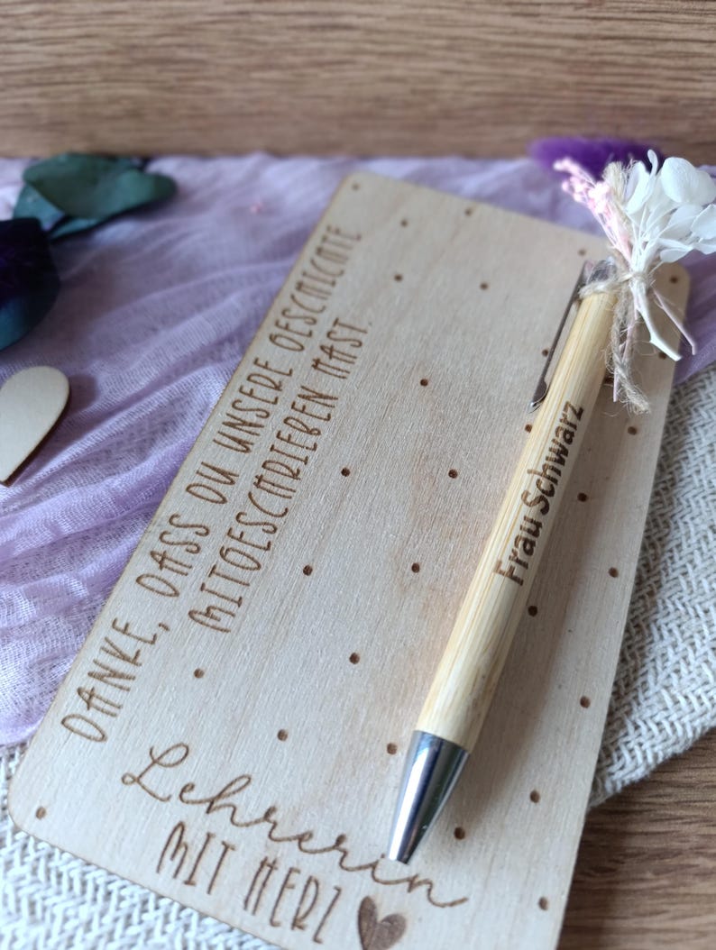 K&ouml;nnte beinhalten: Eine Holzkarte mit graviertem Text und einem Bambusstift. Der Stift ist mit "Frau Schwarz" beschriftet. Auf der Karte stehen die Worte "Danke, dass du unsere geschichte mitschreiben mast" und "Lehrerin mit Herz". Ein kleiner Strau&szlig; wei&szlig;er Blumen und lila Federn ist am Stift befestigt.