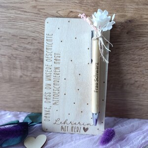 K&ouml;nnte beinhalten: Ein Holznotizblock mit dem Text "DANKE, DASS DU UNSERE GESCHICHTE MITGESCHRIEBEN HAST." und "Lehrerin MIT HERZ" wird angezeigt. Ein Holzstift mit dem Namen "Frau Schwarz" ist mit einer wei&szlig;en Blume und dekorativen Akzenten befestigt. Ein Holzherz ist ebenfalls vorhanden.