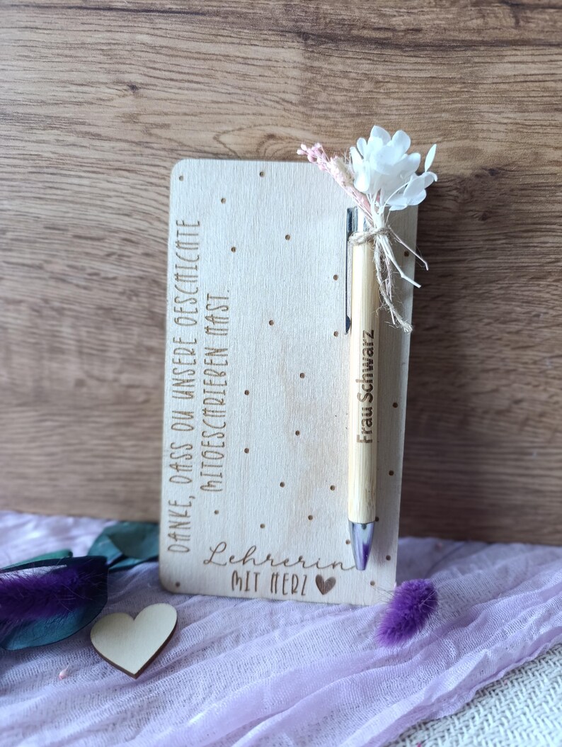 K&ouml;nnte beinhalten: Ein Holznotizblock mit dem Text "DANKE, DASS DU UNSERE GESCHICHTE MITGESCHRIEBEN HAST." und "Lehrerin MIT HERZ" wird angezeigt. Ein Holzstift mit dem Namen "Frau Schwarz" ist mit einer wei&szlig;en Blume und dekorativen Akzenten befestigt. Ein Holzherz ist ebenfalls vorhanden.