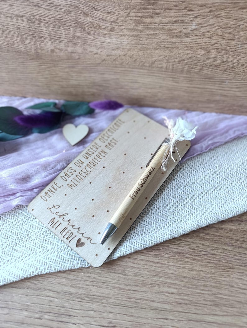 K&ouml;nnte beinhalten: Ein hellbrauner Holzstift und eine Holzkarte mit graviertem Text. Der Stift hat silberne Akzente und eine kleine wei&szlig;e Blume. Die Karte tr&auml;gt den Text "Danke, dass du unsere Geschichte mitschreiben hast. Lehrerin mit Herz" und den Namen "Frau Schwarz".