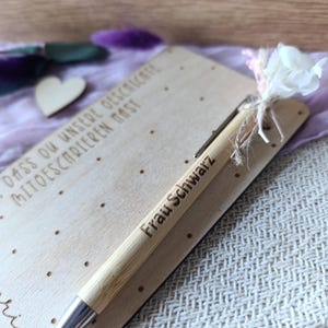 K&ouml;nnte beinhalten: Ein hellbrauner Bambusstift mit dem Text "Frau Schwarz" auf der Seite, der auf einer hellen Holzkarte liegt. Der Stift hat eine silberne Spitze und liegt neben einer kleinen wei&szlig;en Blume und einem hellvioletten Tuch.