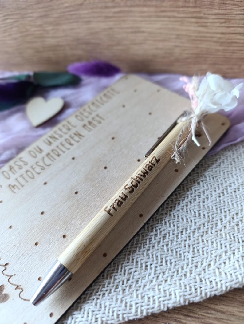 K&ouml;nnte beinhalten: Ein hellbrauner Bambusstift mit dem Text "Frau Schwarz" auf der Seite, der auf einer hellen Holzkarte liegt. Der Stift hat eine silberne Spitze und liegt neben einer kleinen wei&szlig;en Blume und einem hellvioletten Tuch.