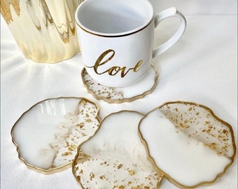 May include: Set of four white resin coasters with gold leaf accents and a gold rim. The coasters are shaped like irregular circles and have a slightly raised edge. A white ceramic mug with a gold rim and the word "love" written in gold script is sitting on one of the coasters.