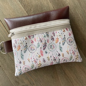 May include: A small, rectangular pouch with a brown leather top and a white fabric bottom. The fabric is patterned with colorful dreamcatchers and feathers. A beige zipper and a silver clasp are visible.