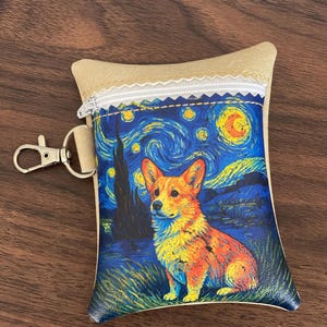 May include: A rectangular pouch with a beige top section and a zipper closure. The main body features a colorful print of a Corgi dog set against a swirling blue and yellow night sky background. A silver metal lobster clasp is attached to the side.