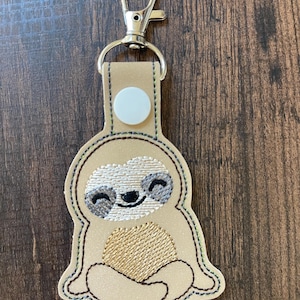 May include: A tan sloth keychain with a silver clasp. The sloth has a smiling face with embroidered details in shades of gray and tan. The sloth's belly has a textured, embroidered design. The keychain is attached to a tan leather-like backing.