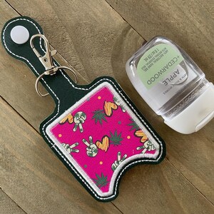 May include: A green fabric keychain with a pink and yellow peace sign and marijuana leaf pattern. The keychain has a loop for attaching to a bag or belt. A clear plastic bottle of hand sanitizer is attached to the keychain. The bottle has a white label with the text "Apple Cedarwood" and "Bath & Body Works".