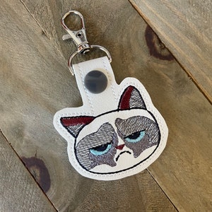 May include: White leather keychain with a grumpy cat face embroidered on it. The cat has gray fur, blue eyes, and a red collar. The keychain has a silver metal ring.