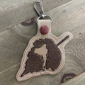 May include: A brown and tan leather keychain with a brown glitter silhouette of a person holding a sword. The keychain has a silver clip and a brown button.