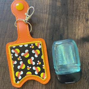 May include: An orange hand sanitizer holder with a candy corn pattern and a matching sanitizer bottle. The holder has a yellow button and a silver clasp. The sanitizer bottle is clear with blue liquid.