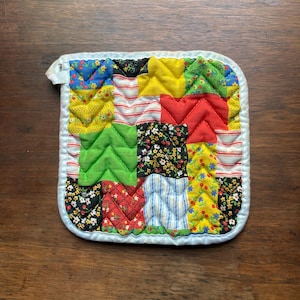 May include: A square quilted potholder featuring a patchwork design of various fabric squares in bright colors including yellow, red, green, blue, and black with floral, cherry, and striped patterns. It has a white fabric border and a small loop.