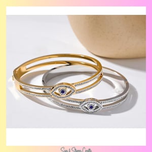 May include: Two gold and silver bracelets with a blue evil eye charm. The bracelets are adorned with clear rhinestones.