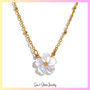 Könnte beinhalten: Eine goldfarbene Halskette mit einem weißen Blumenanhänger. Die Blume hat mehrere Blütenblätter und eine goldfarbene Mitte. Die Kette ist ebenfalls goldfarben und hat kleine, runde Perlen. Der Text "Sun & Storm Jewelry" steht unten.