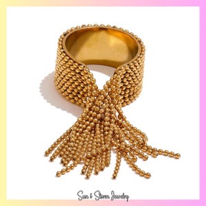 May include: Gold-coloured cuff bracelet with a beaded design and dangling fringe. The bracelet features a wide band covered in small, round beads. The fringe is made of strands of similar beads. The jewellery is set against a white background.
