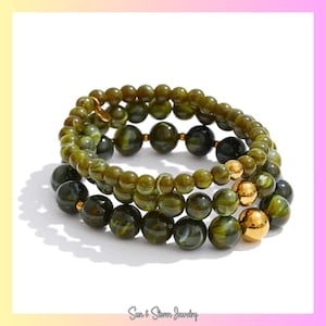 May include: Three stacked bracelets featuring olive green and black beads, accented with gold-colored beads. The bracelets are made of round beads of varying sizes. The text "Sun & Storm Jewelry" is at the bottom.