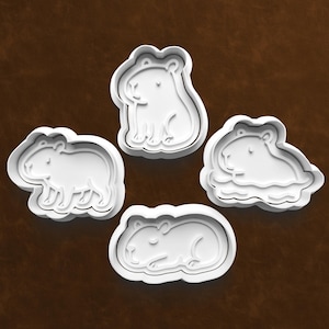May include: Four white plastic capybara-shaped cookie cutters. Each cutter depicts a different pose of the animal: standing, sitting, swimming, and lying down. The cutters are on a brown background.