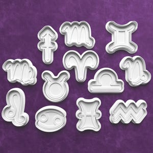 May include: A set of white plastic cookie cutters, each representing a zodiac sign. The cutters are displayed on a purple surface, with each sign clearly visible. The shapes include Aries, Taurus, Gemini, Cancer, Leo, Virgo, Libra, Scorpio, Sagittarius, Capricorn, Aquarius, and Pisces.
