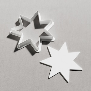May include: Two white star-shaped cookie cutters. One is a solid star, and the other is a star-shaped cutter with a raised edge. The cutters are on a grey surface.