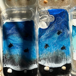 May include: Clear phone cases with ocean-themed resin art. The designs feature dark blue and white waves, black sand, and small sea turtle silhouettes. Some cases include seashells.