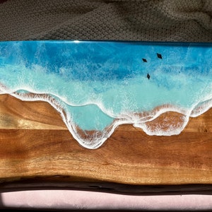 Puede incluir: Una tabla de servir de madera con un diseño de resina epoxi azul y blanca que se asemeja a una escena oceánica con olas rompiendo en la costa. Hay tres siluetas negras de pájaros volando sobre el agua.